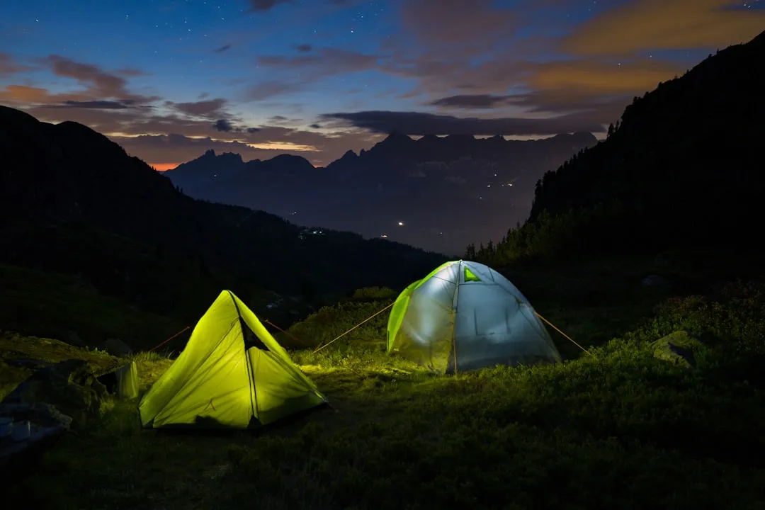 Two glowing tents on a mountain meadow at dusk with mountain silhouettes behind