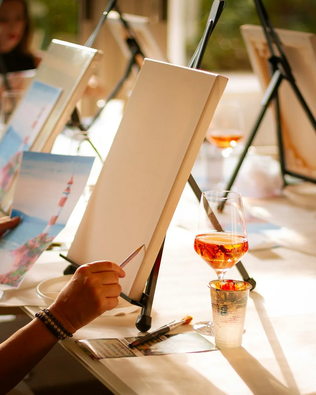 Hand holding a paintbrush at an easel with a wine glass during a paint and sip class
