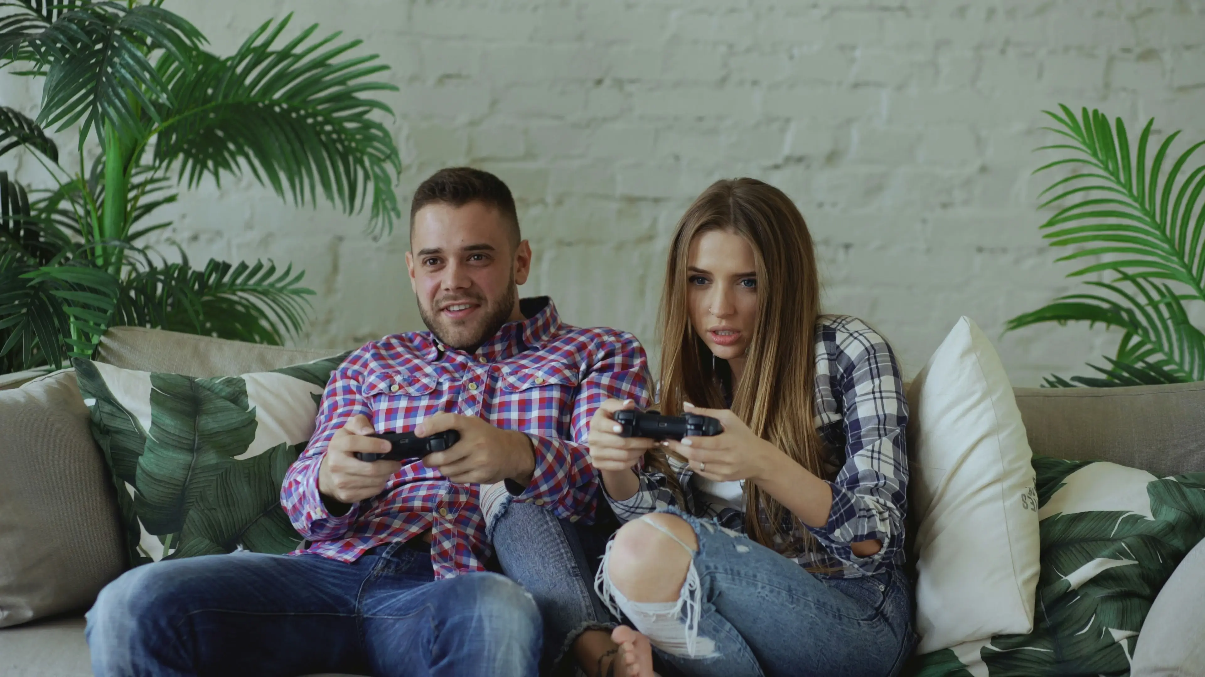 Couple sitting on couch with controllers, woman focused on gameplay while man relaxes