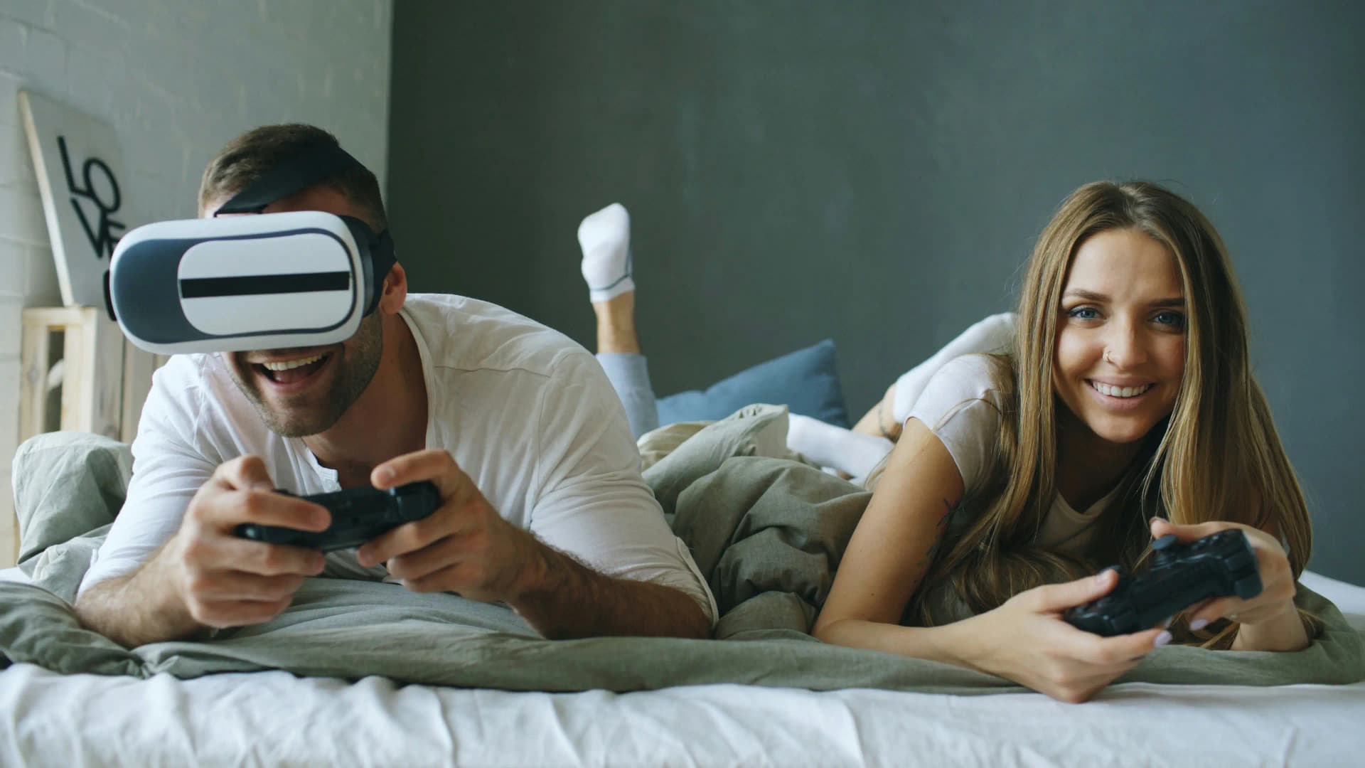 Couple relaxed on bed playing video games, one wearing VR headset