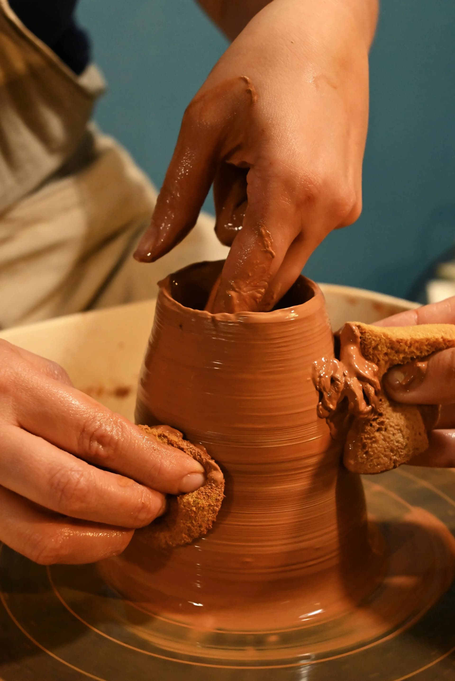 Hands covered in wet clay shaping a pot on the pottery wheel